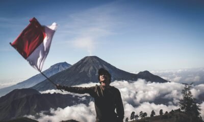 A hiker waves the Indonesian flag atop a scenic mountain, surrounded by clouds.