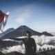 A hiker waves the Indonesian flag atop a scenic mountain, surrounded by clouds.